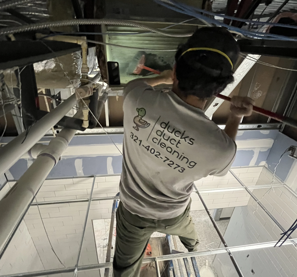 Worker wearing a ducks duct cleaning shirt cleaning the filter of an air duct in the ceiling with a broom.